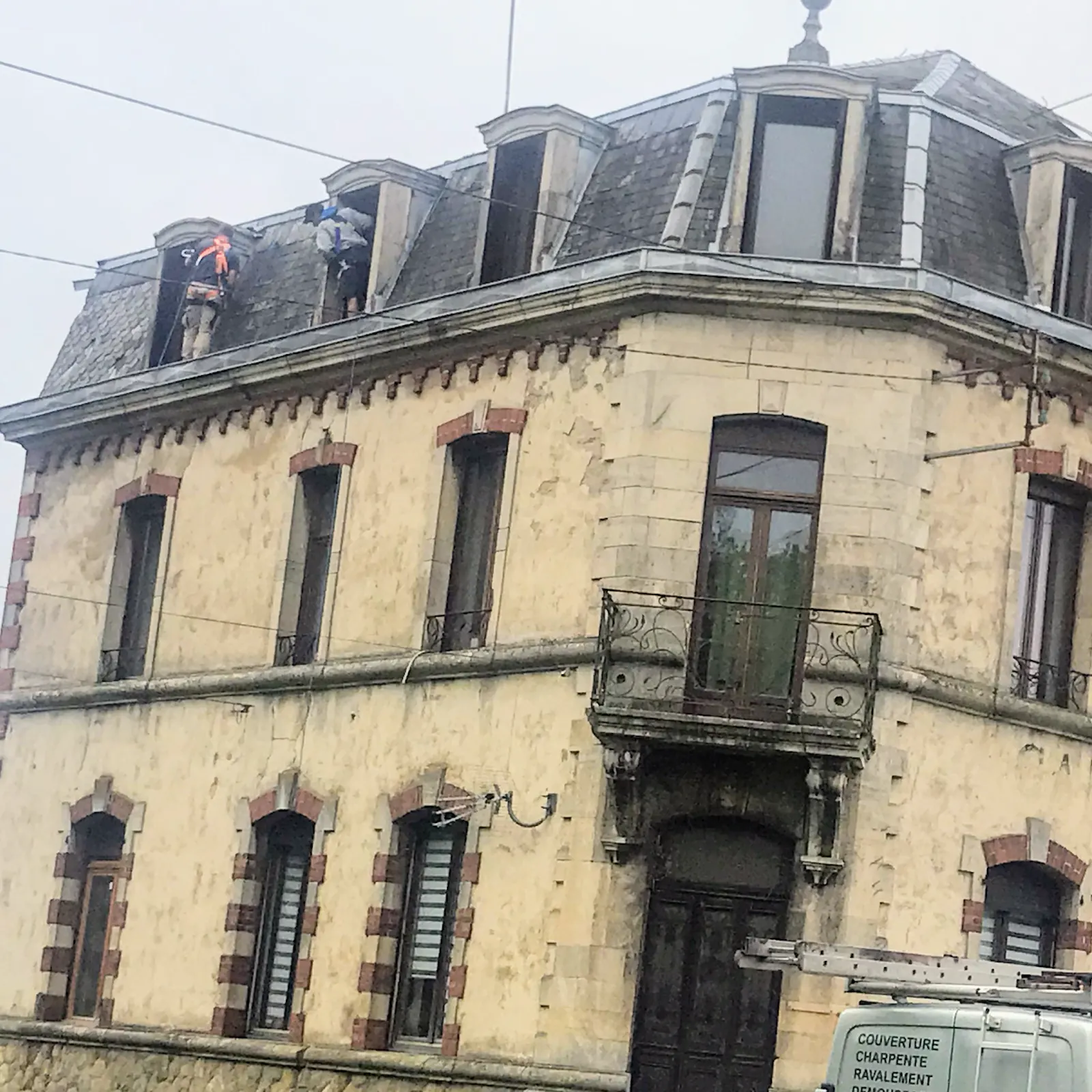 Deux couvreurs harnachés sur le toit à la Mansart d’un immeuble ancien, camionnette garée devant
