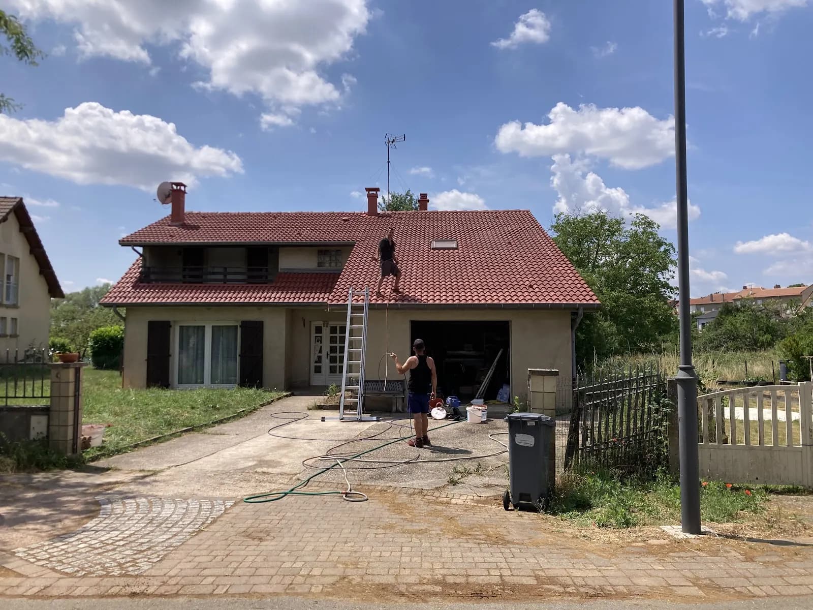 Couvreur sur une toiture en tuiles rouges, un second au pied de l’échelle avec le tuyau d’eau
