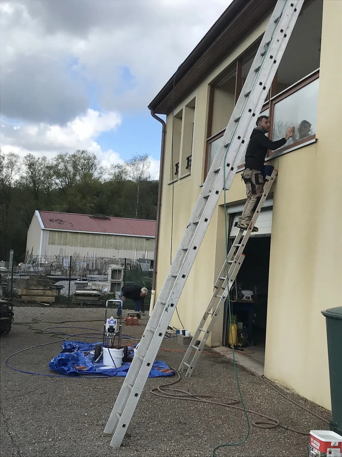 Ouvrier en haut d’une échelle contre une façade, bâche et nettoyeur au pied du mur
